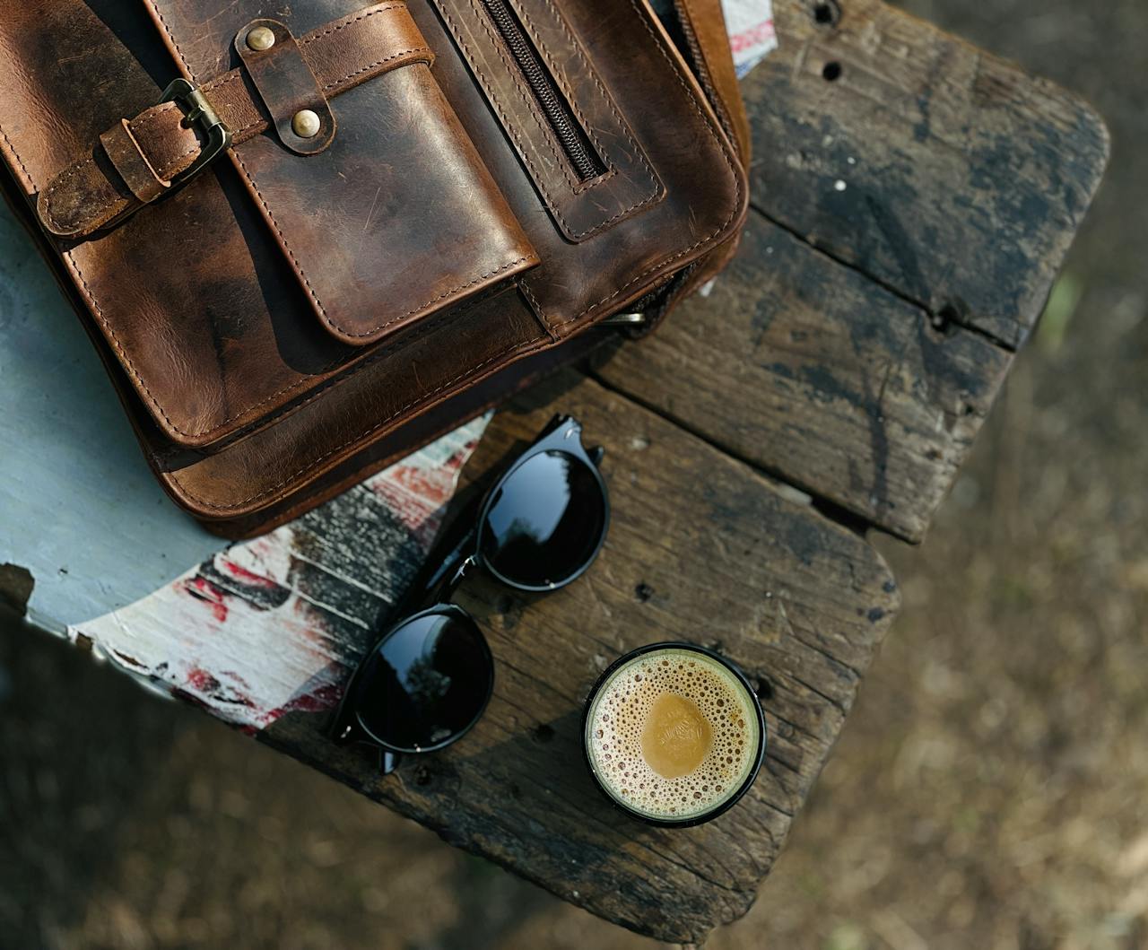 Top view of a leather bag, sunglasses, and coffee on a rustic wooden table outdoors, evoking a vintage lifestyle.