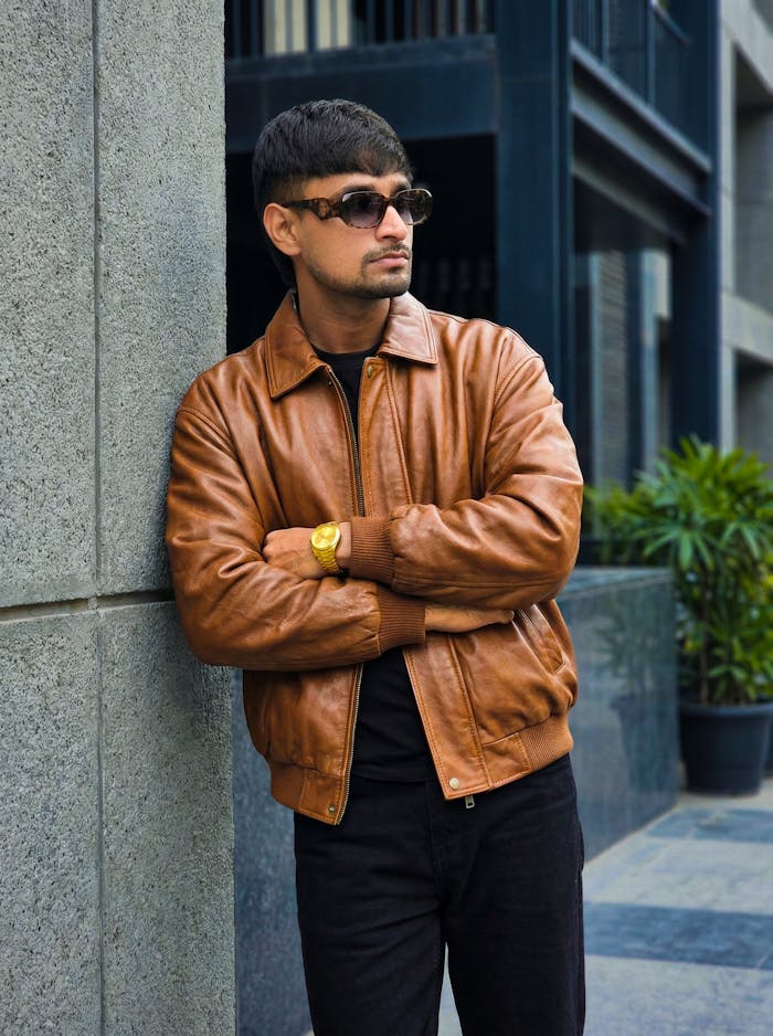 services-02 Man wearing sunglasses and a brown leather jacket, leaning against an urban wall, exuding confidence.